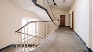 A long, narrow hallway with a staircase on the left. The hallway has a wooden door at the end and a window on the left side. - Plac Konstytucji Studio (Warsaw)