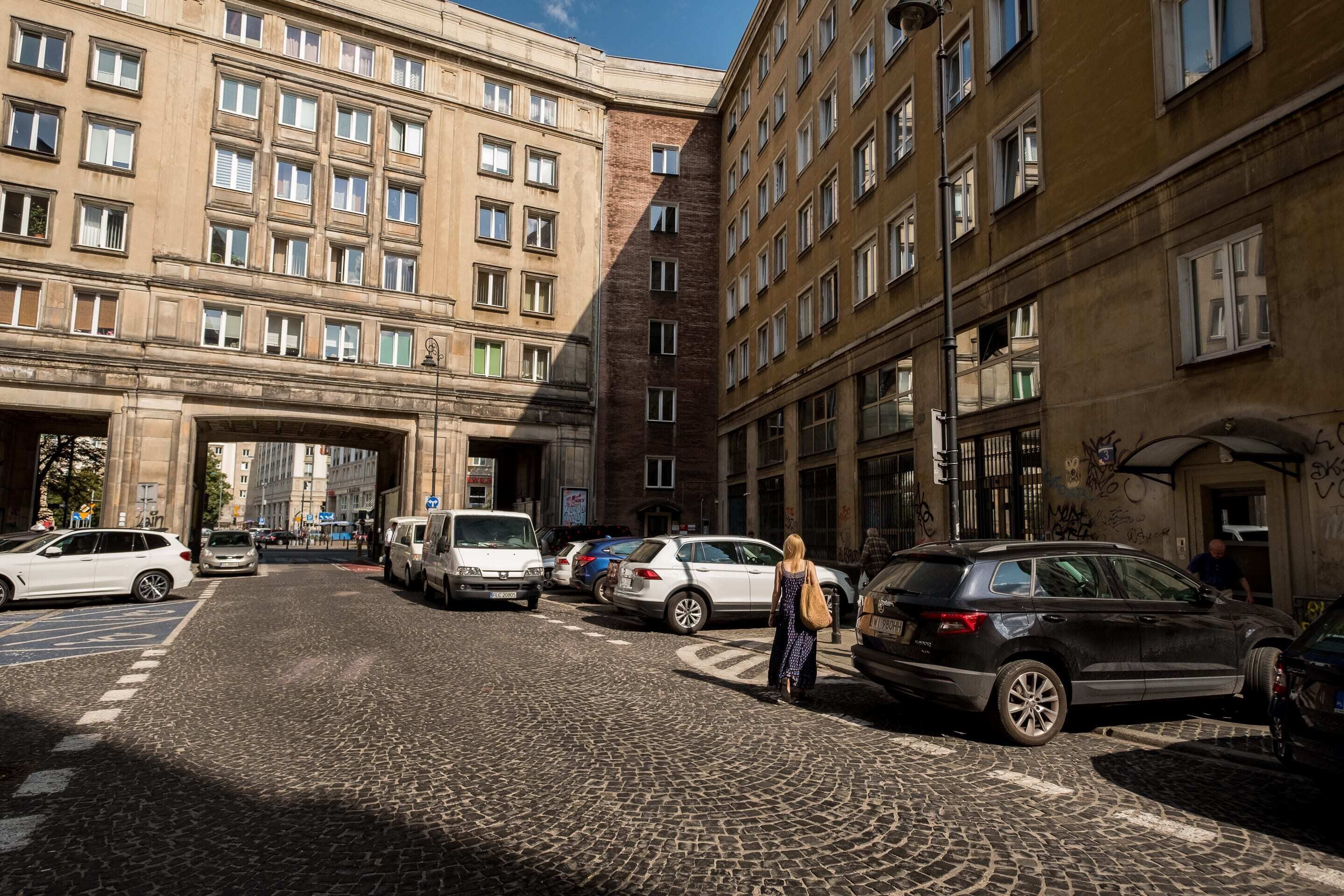 Una calle adoquinada con coches aparcados y un edificio grande con varias ventanas y balcones.