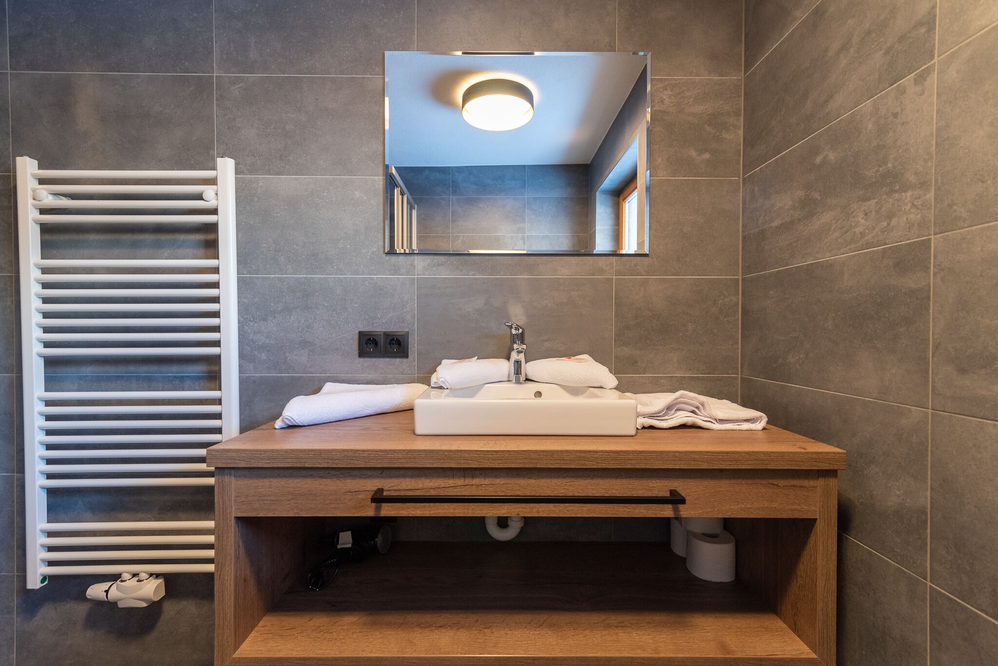 A modern bathroom with a wooden vanity, a white sink, and a mirror above it. There is a towel rack to the left and a towel on the sink.