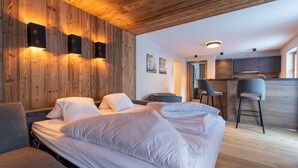Quarto de hotel moderno com painéis de madeira, uma cama grande, duas cadeiras, uma mesinha e uma kitchenette ao fundo.