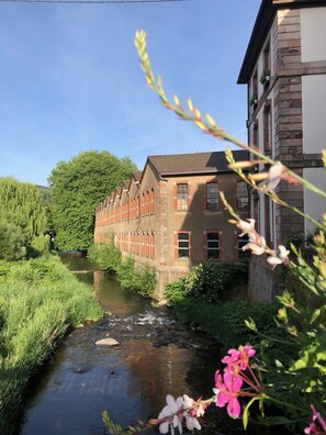 A building with a long, narrow structure, a river in front, and greenery around. - Satin Take advantage of this comfortable and refined apartment for 2 people. (Masevaux)