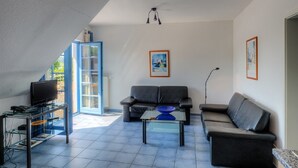 Una sala moderna con un sofá, una mesa de centro y un televisor. Se ve un ventanal con vistas a zonas verdes.