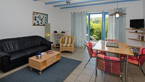 Sala de estar con un sofá de cuero negro, una mesa de centro de madera, una silla de mimbre, una mesa de comedor con sillas rojas, un televisor en un estante y una ventana con cortinas.