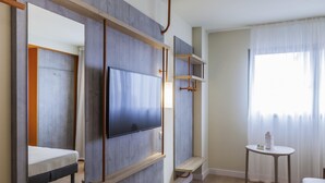 Una habitación de hotel moderna con una televisión de pantalla plana montada en un panel de madera, una mesita con una botella y una cama con colchón y almohadas.