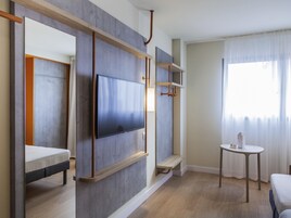 Una habitación de hotel moderna con una televisión de pantalla plana montada en un panel de madera, una mesita con una botella y una cama con colchón y almohadas.