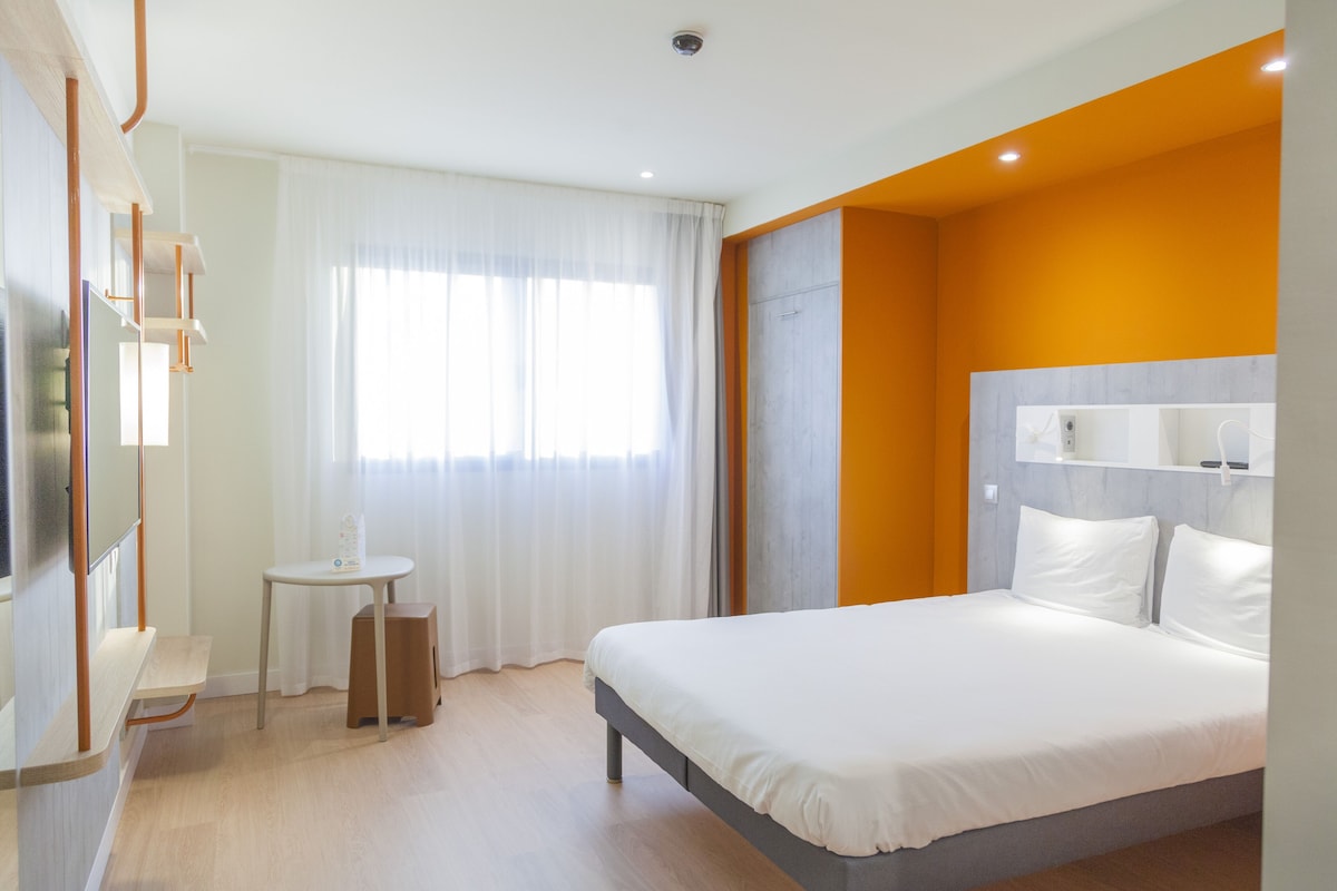 Una habitación de hotel moderna con una cama, una mesita y un ventanal con cortinas.