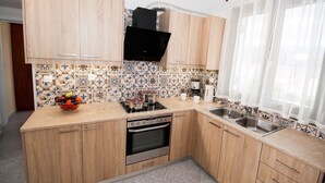 A kitchen with wooden cabinets, a tiled backsplash, a stove, a sink, and a window with sheer curtains. - Charming Apartment in Kefalonia Island (Kefalonia)