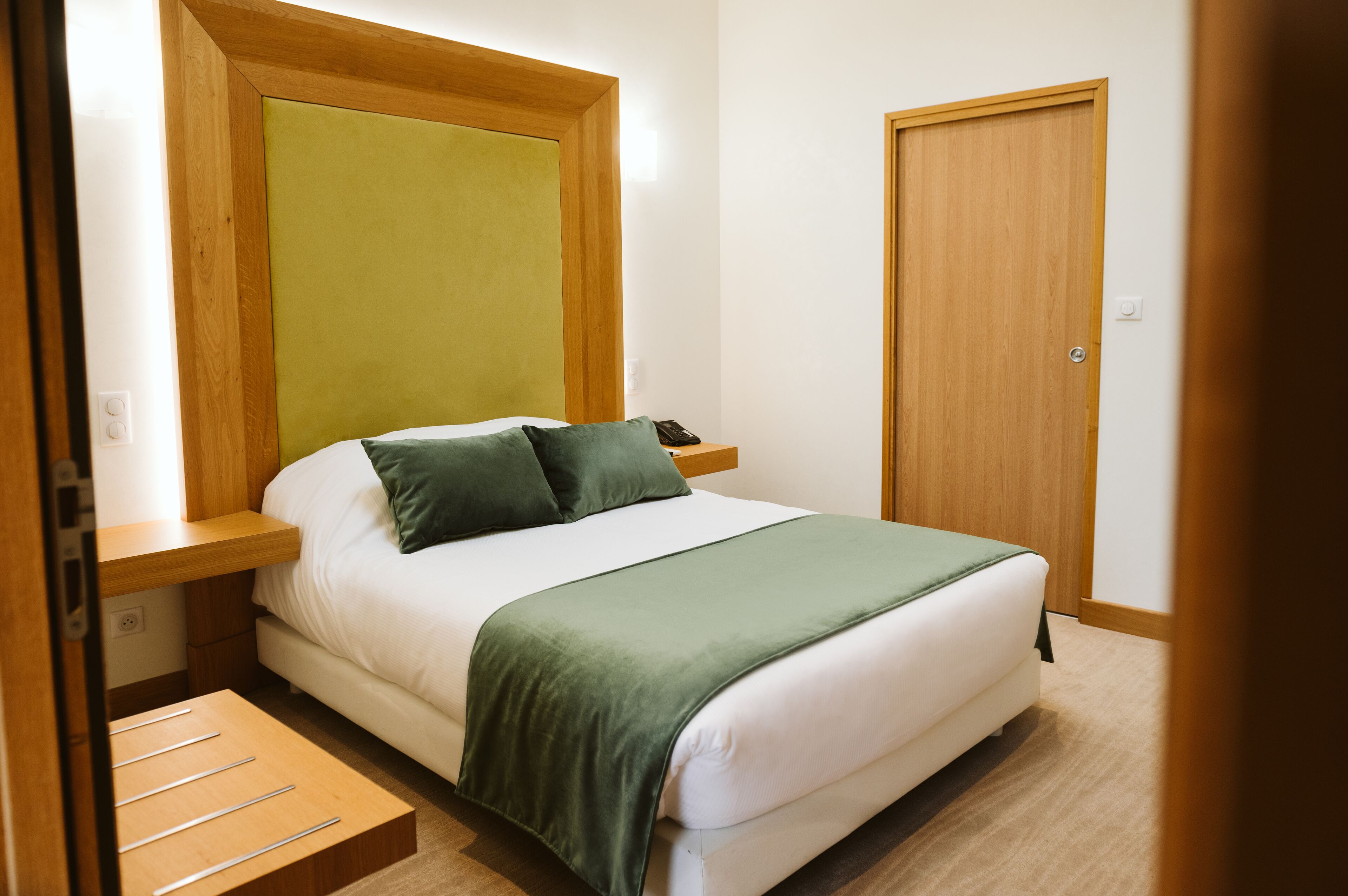 A neatly made bed with a green blanket, wooden headboard, and bedside tables with a phone and lamp.