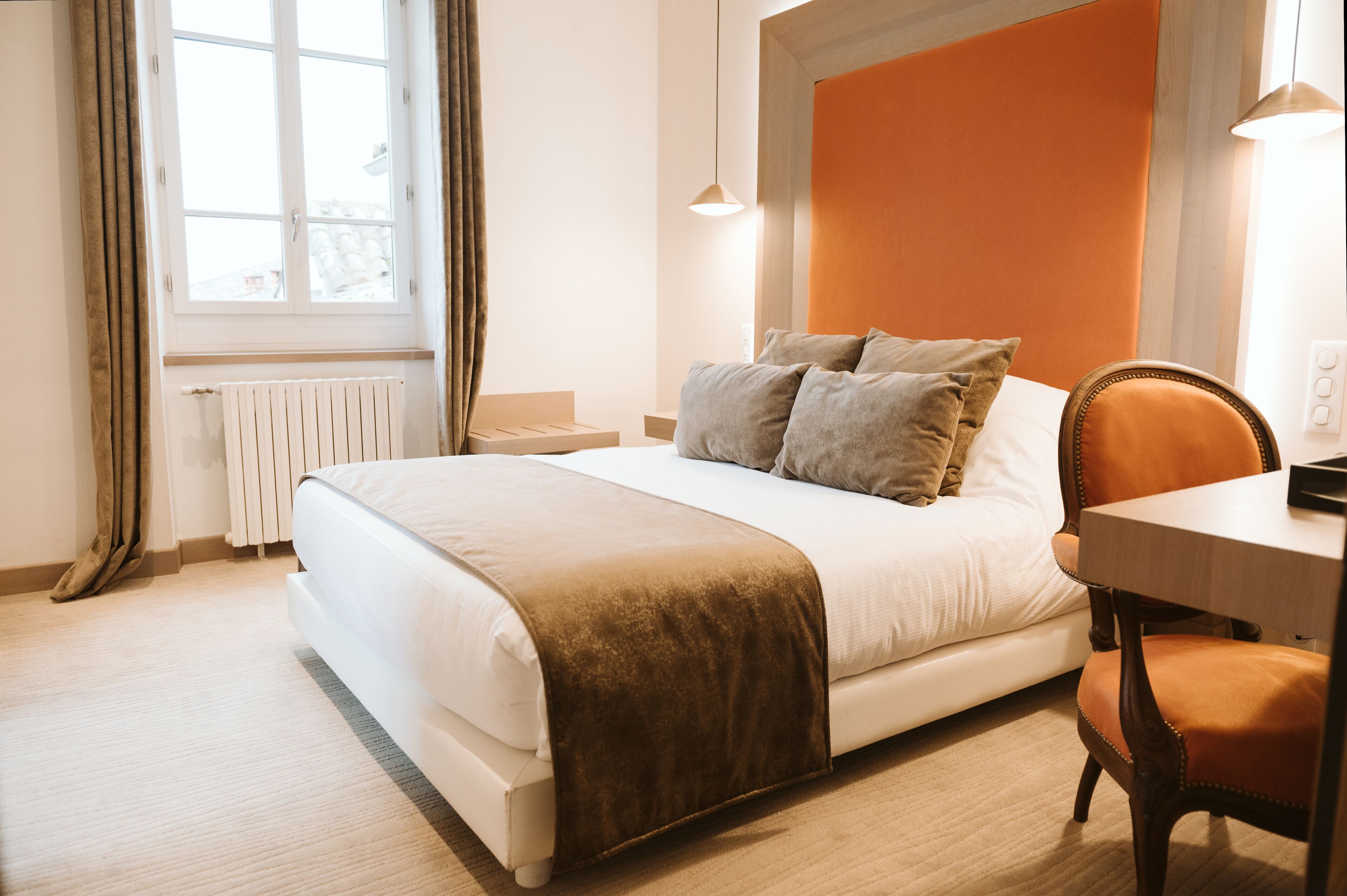 A neatly made bed with a brown throw blanket, a wooden nightstand, a chair, and a window with curtains.