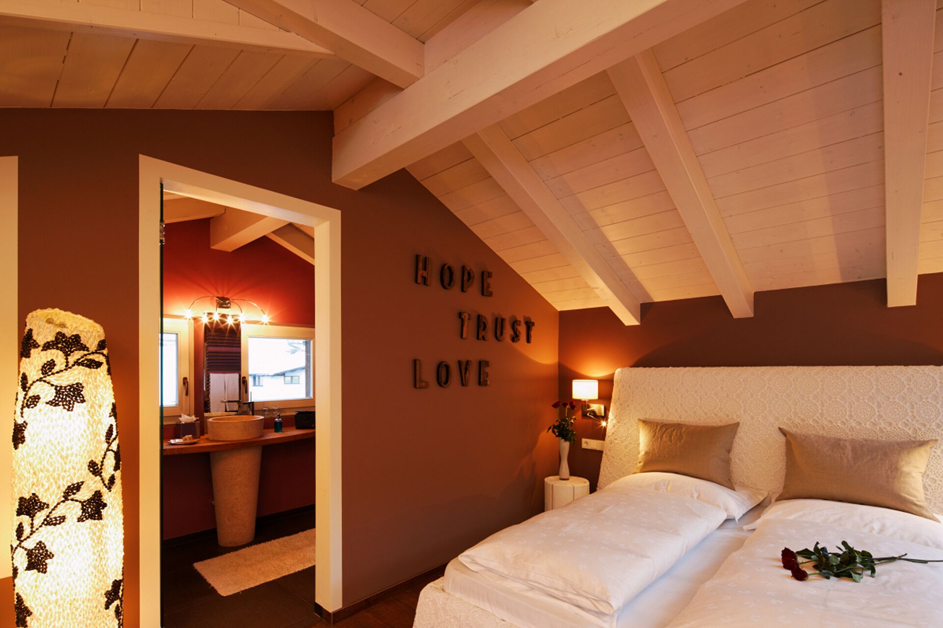 A bedroom with a bed, a nightstand, a wall with 'HOPE TRUST LOVE' text, and a bathroom visible through an open door.