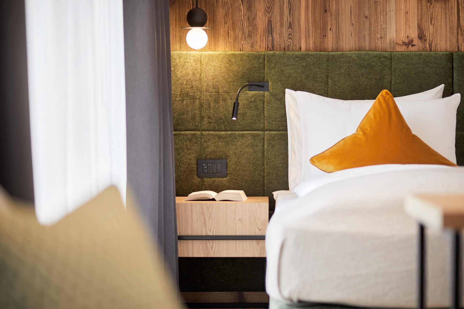 A neatly made bed with white and yellow pillows, a bedside table with a book, and a wall-mounted light fixture.