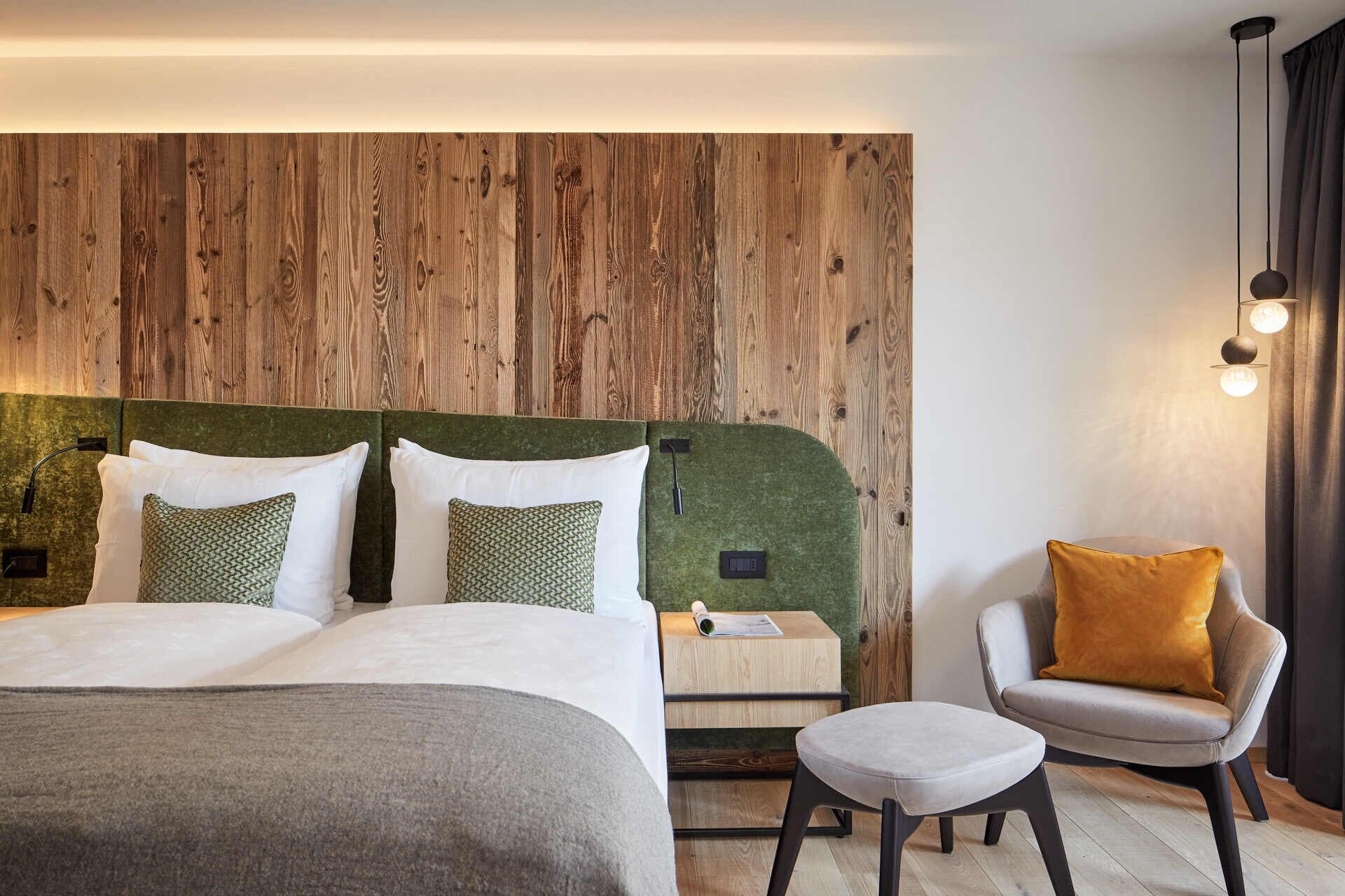 Una habitación de hotel moderna con cabecera de madera, cama con almohadas blancas y verdes, mesita de noche con lámpara, sillón gris con cojín amarillo y un pequeño otoman.