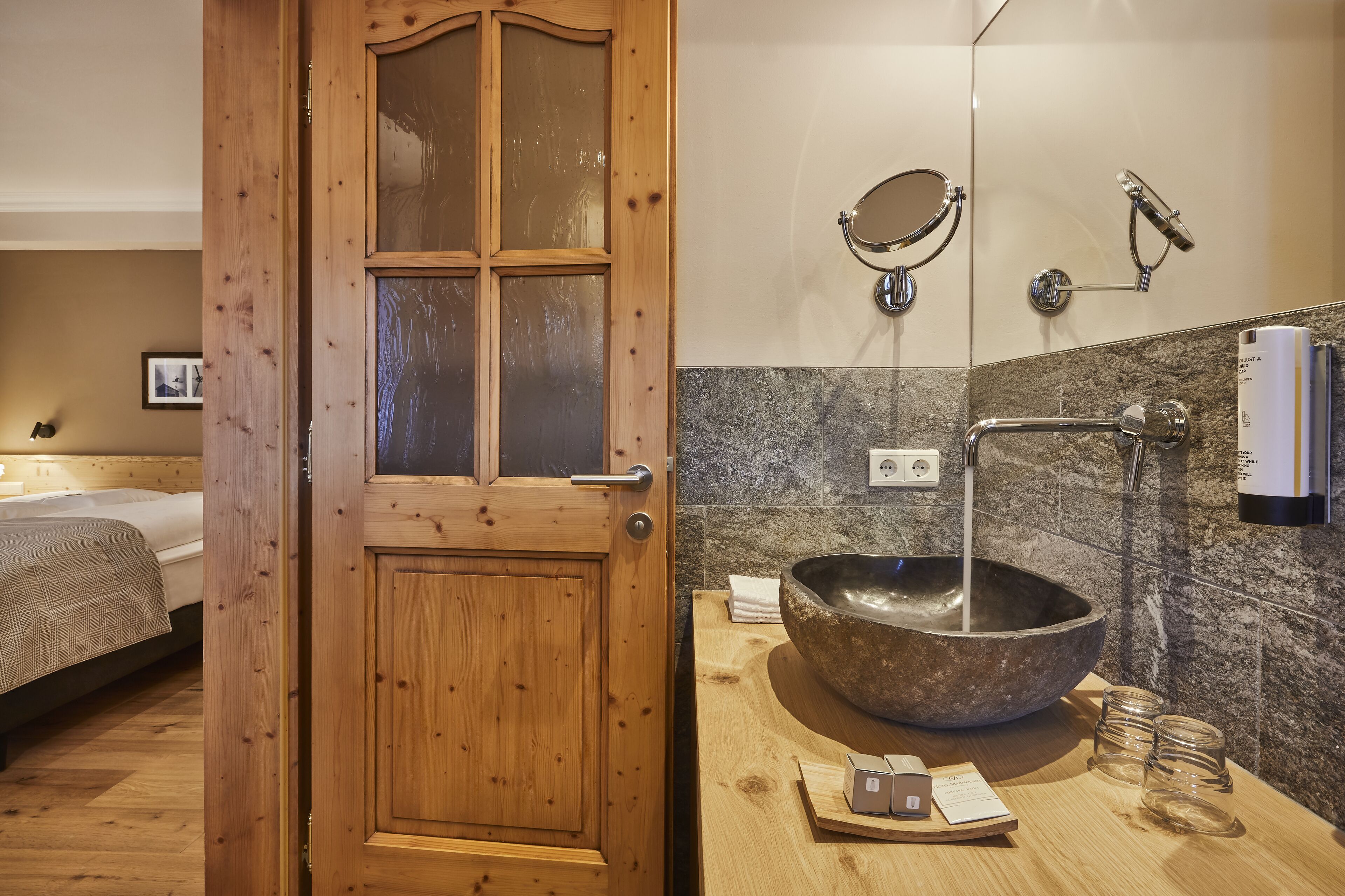 A wooden door with glass panels, a bathroom with a large stone sink, a mirror, a handheld showerhead, and a towel dispenser.