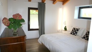 A bedroom with a wooden ceiling, a bed with white bedding, a dresser, a vase with flowers, a window with curtains, and a small table with a plant. - Civico 1A1 (Santa Maria di Sala)