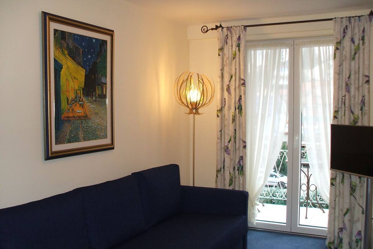 Une pièce avec un canapé bleu foncé, un tableau encadré représentant une scène nocturne, une lampe sur pied et un balcon avec des rideaux blancs.