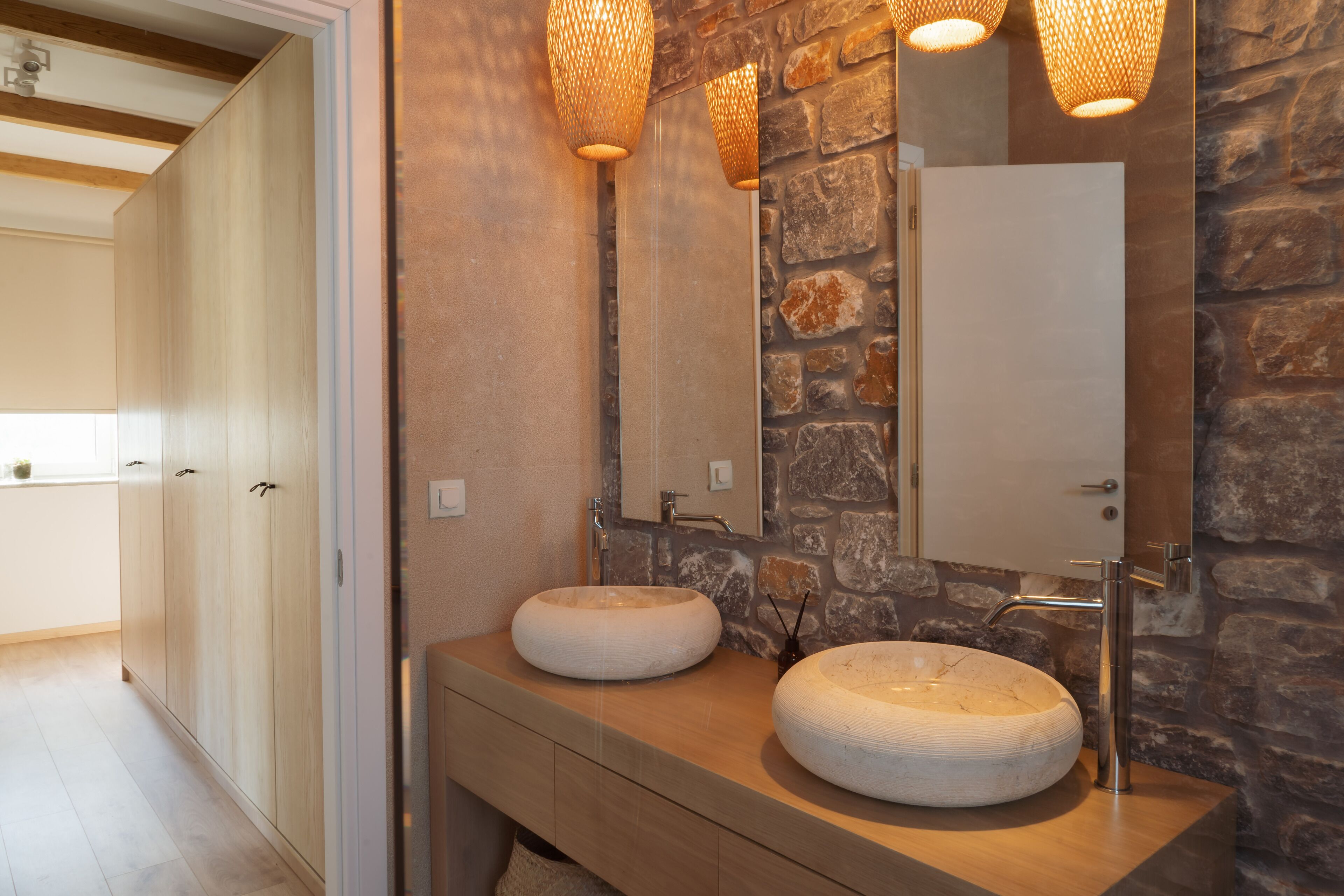 Un baño con dos lavamanos redondos de cerámica blanca, un mueble de tocador de madera, una pared de piedra y un espejo con un gabinete de madera encima.