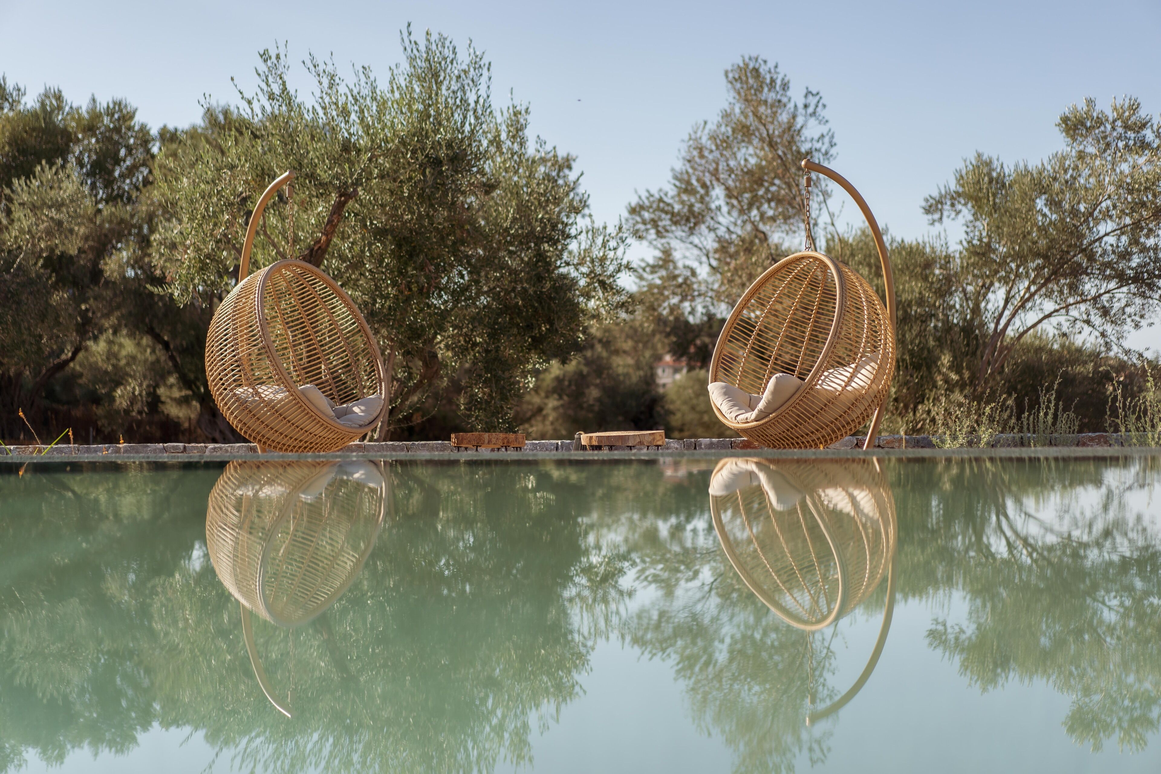 Dos sillas de mimbre junto a una piscina con reflejo transparente, rodeadas de árboles.