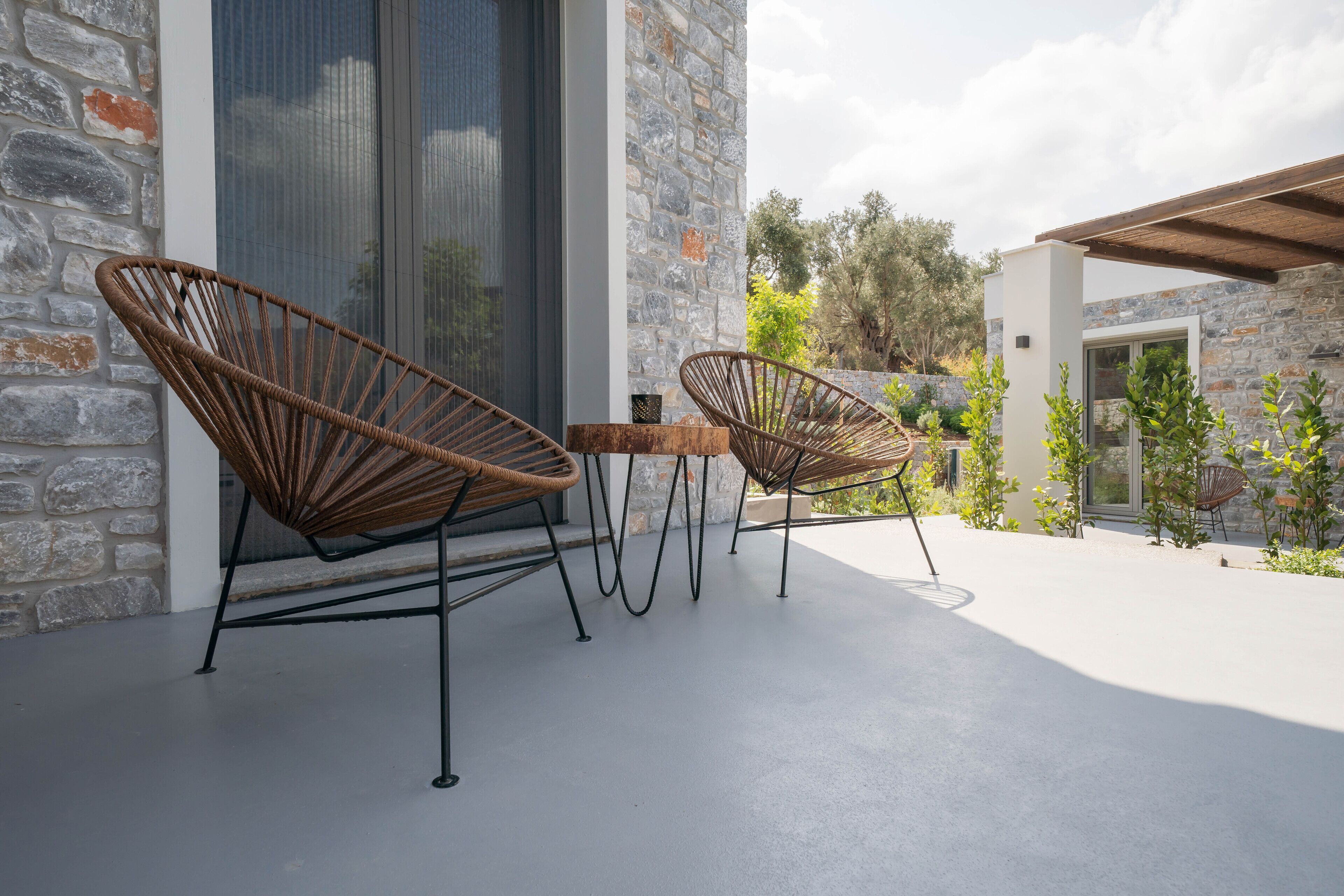 Un patio con dos sillas de mimbre y una pequeña mesa, frente a una pared de piedra y un portón de cristal.