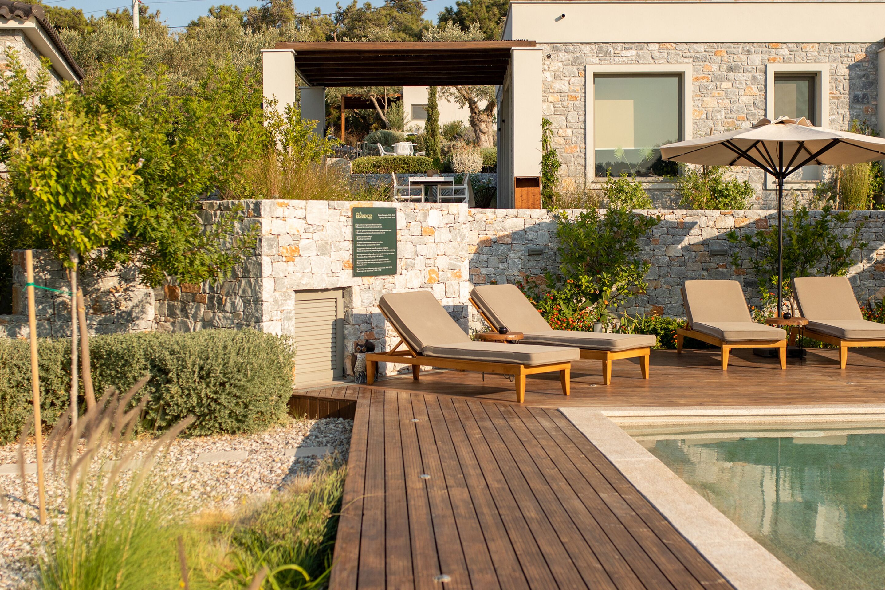 Zona junto a la piscina con sillones de descanso y una sombrilla, rodeada de muros de piedra y vegetación.