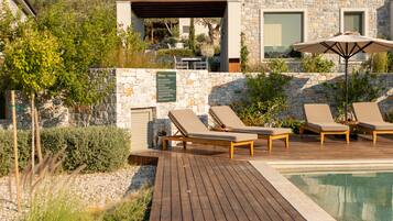 Zona junto a la piscina con sillones de descanso y una sombrilla, rodeada de muros de piedra y vegetación.