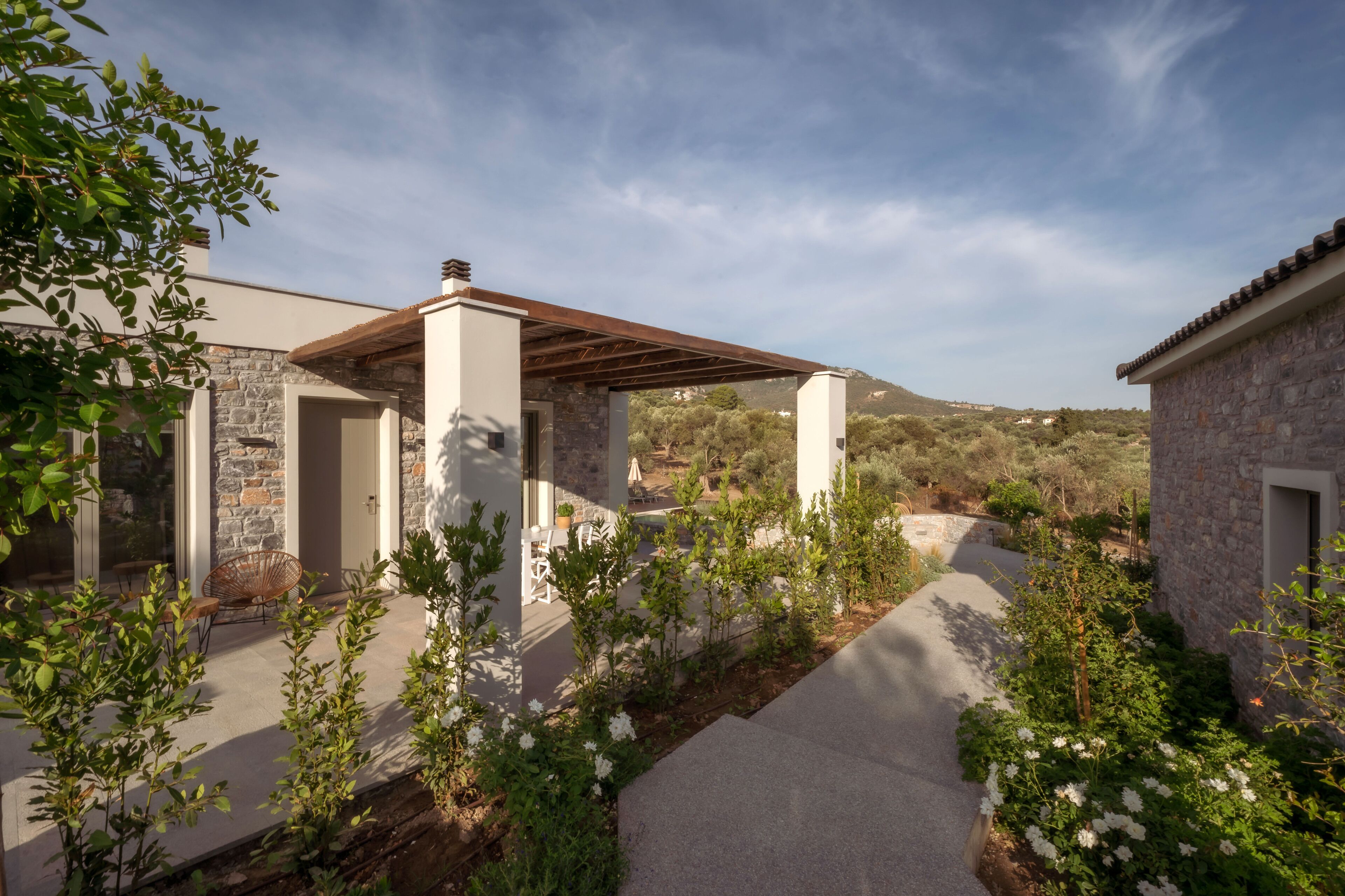 Una casa moderna amb façana de pedra, un porxo cobert i un jardí amb diverses plantes i flors.