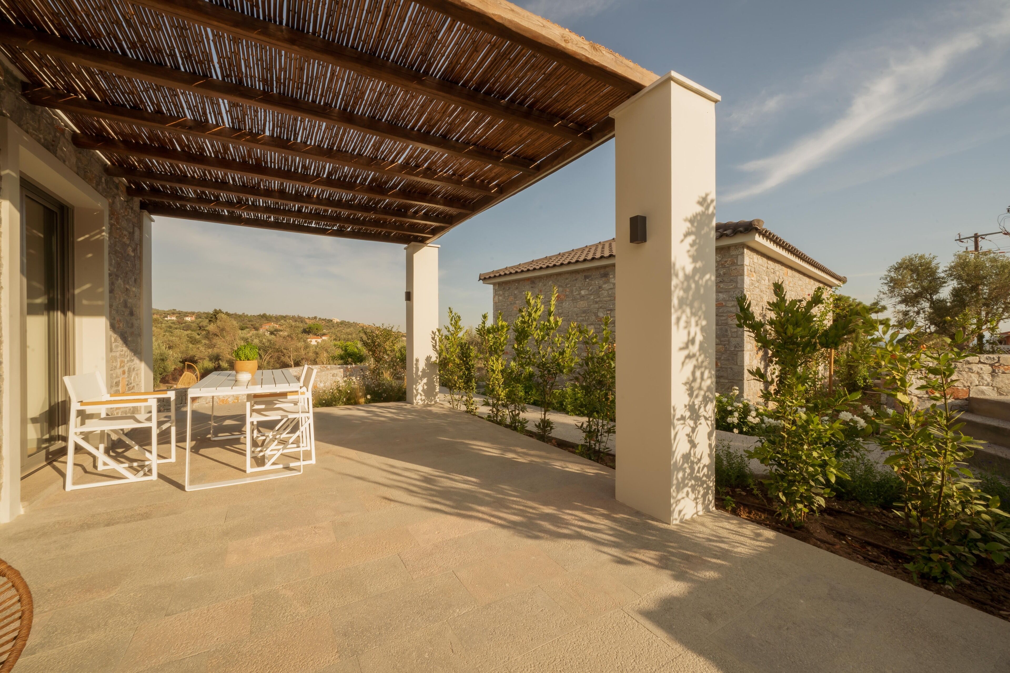 An outdoor patio with a pergola, white furniture, and a view of a stone building.