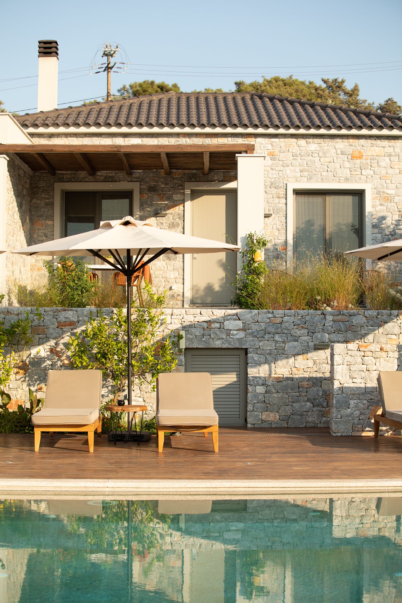 A poolside area with lounge chairs and an umbrella, a stone wall, and a building with a tiled roof.