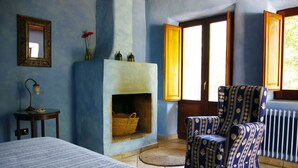 Una habitación con cama, un sillón estampado, chimenea, espejo y ventana con contraventanas abiertas.