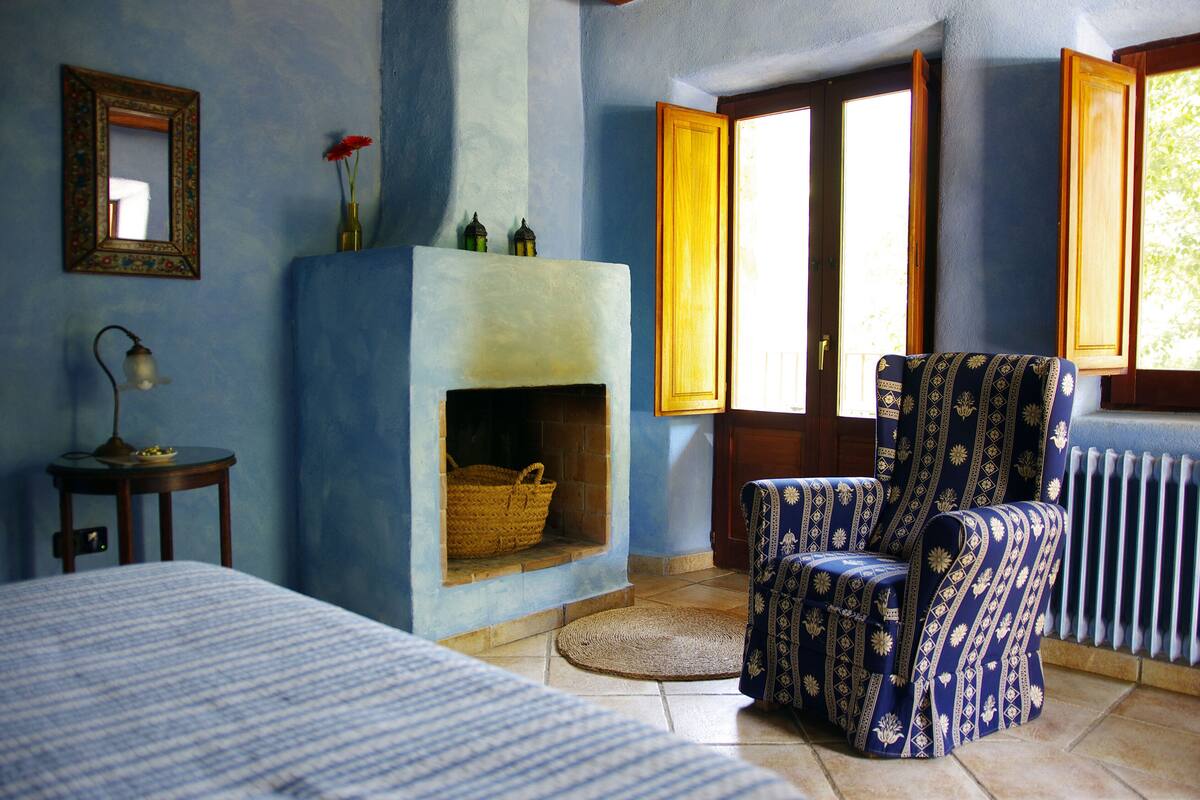 Una habitación con cama, un sillón estampado, chimenea, espejo y ventana con contraventanas abiertas.