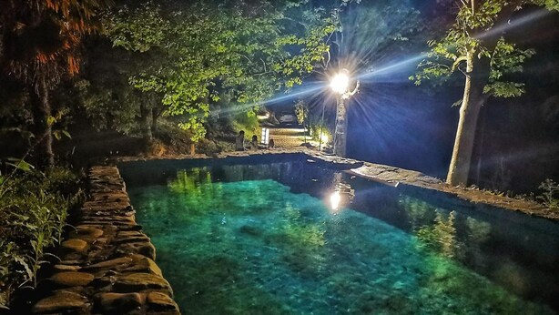 Una piscina cristalina rodeada de muros de piedra y árboles, con una fuente de luz al fondo.