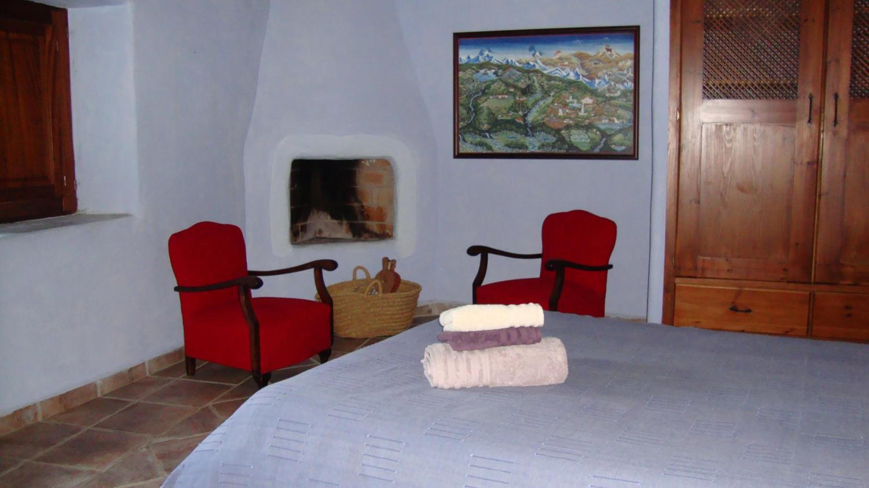 A room with a bed, two red chairs, a fireplace, and a painting on the wall.