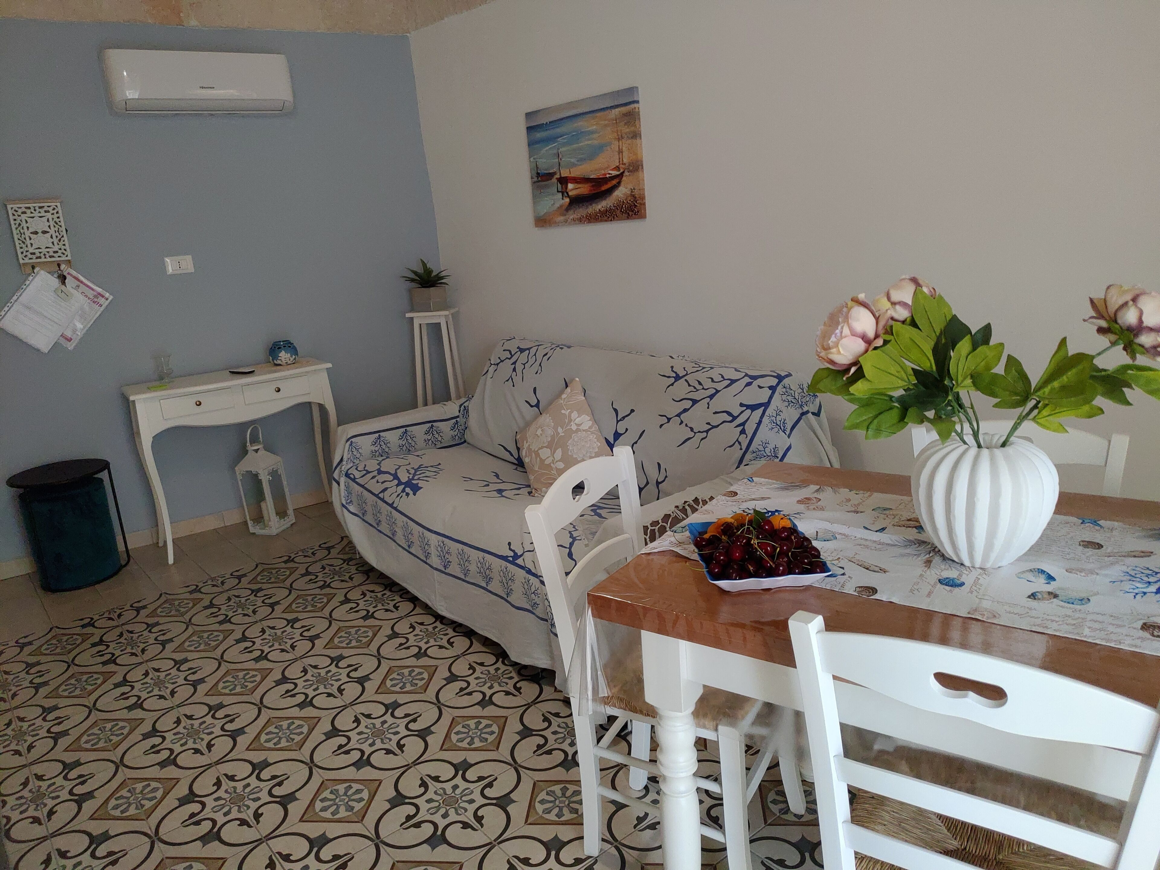 A cozy living room with a patterned floor, a sofa with a tree design, a dining table with a vase of flowers, and a painting on the wall.