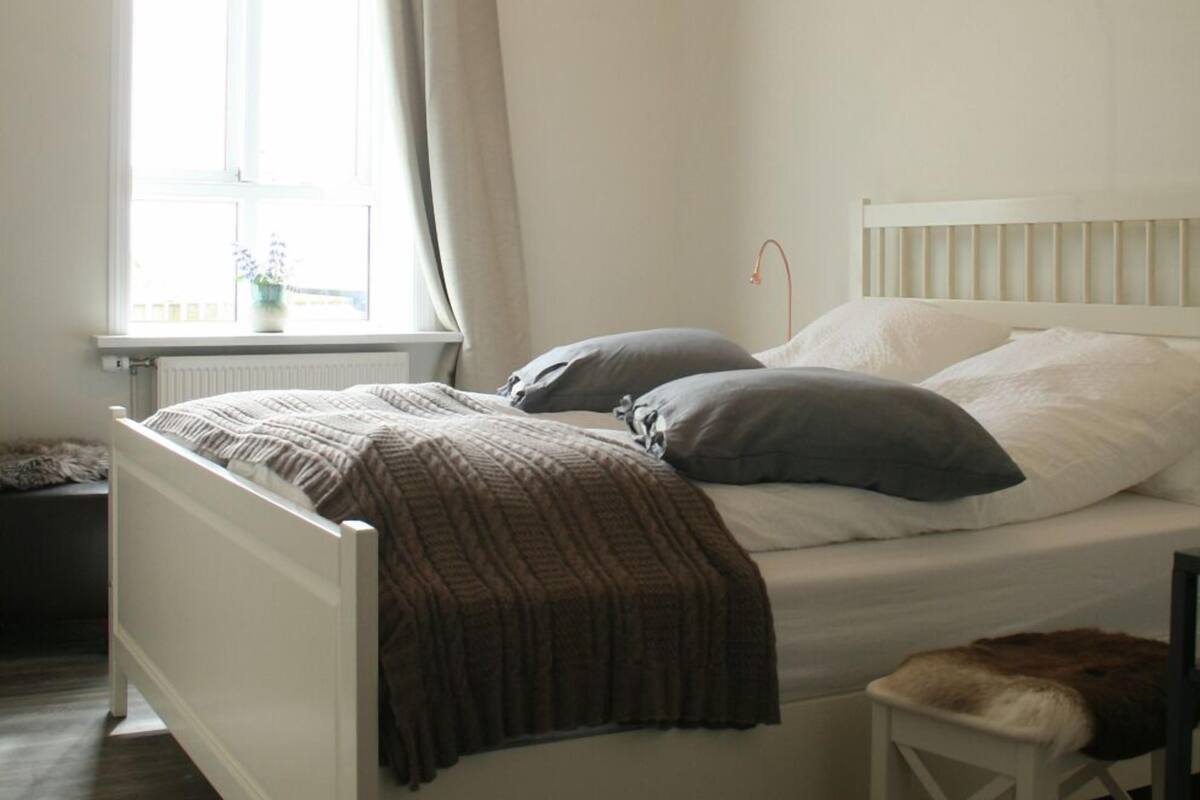 A bedroom with a white bed, a wooden floor, a window with curtains, and a geometric light fixture.