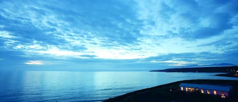 A coastal view with a house overlooking the sea, a clear sky, and distant mountains.