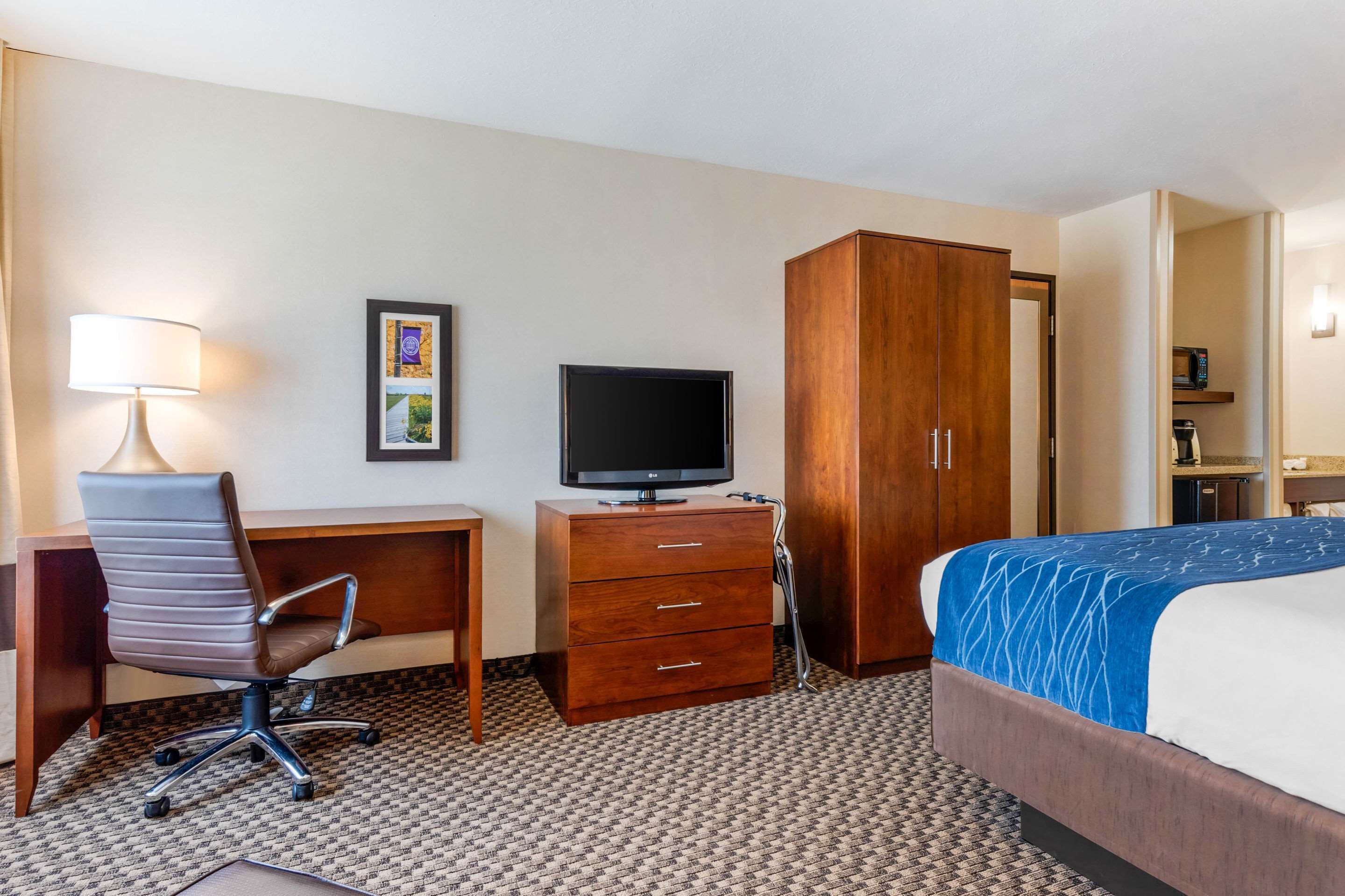 A hotel room with a bed, a desk with a chair, a television, a wardrobe, and a small kitchenette.