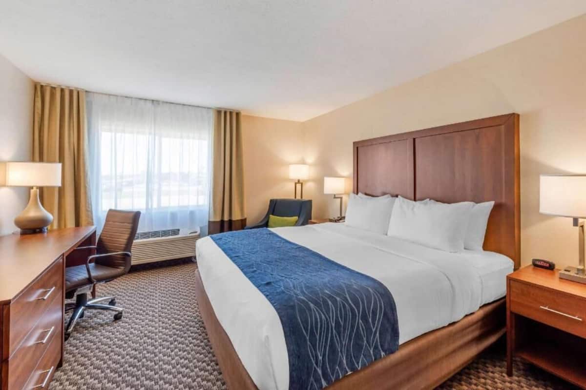 A hotel room with a bed, desk, chair, lamps, and a window with curtains.