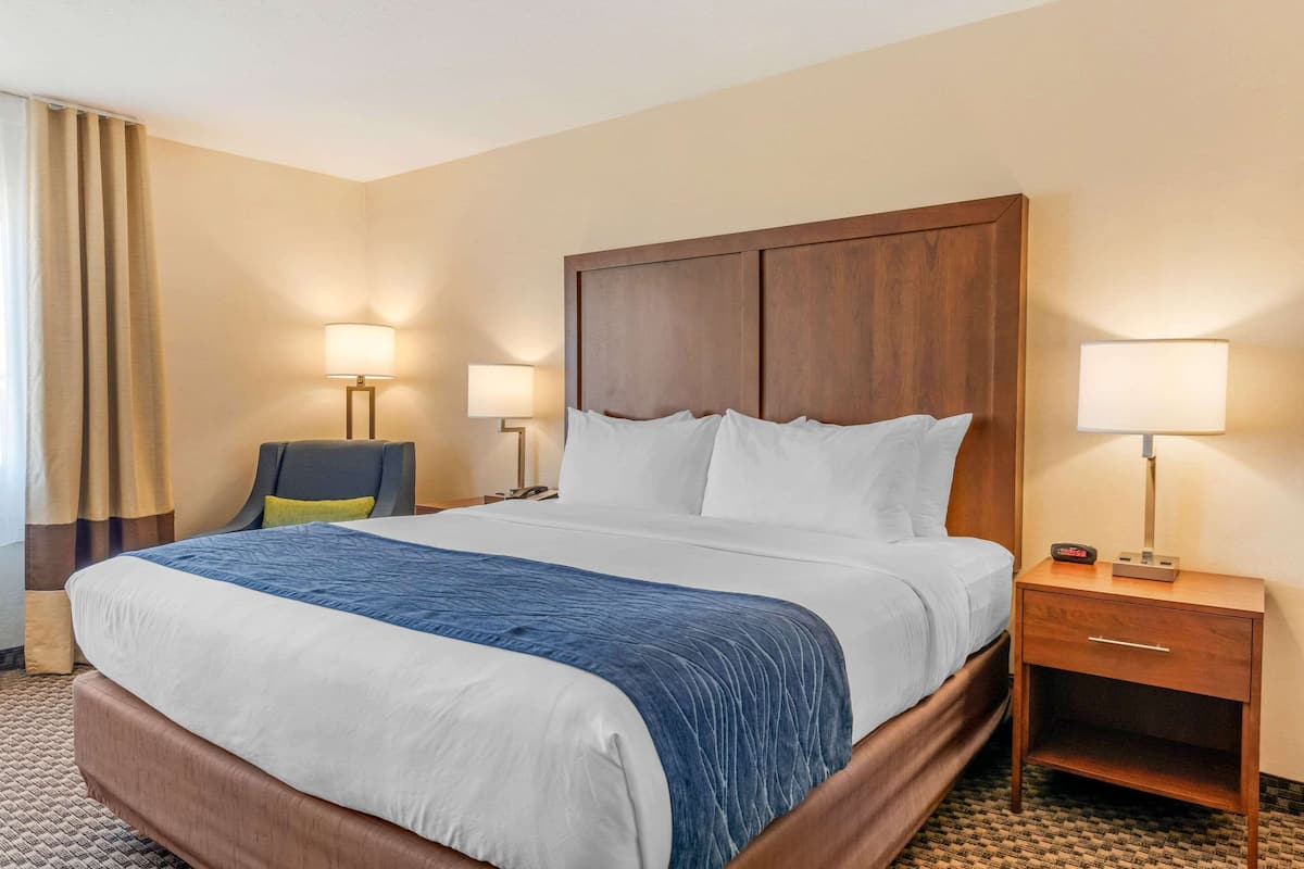 A hotel room with a large bed, a wooden nightstand with a lamp, and a chair.