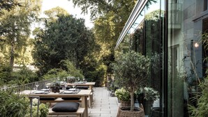 Een terras met houten tafels en banken, omgeven door potplanten en bomen.