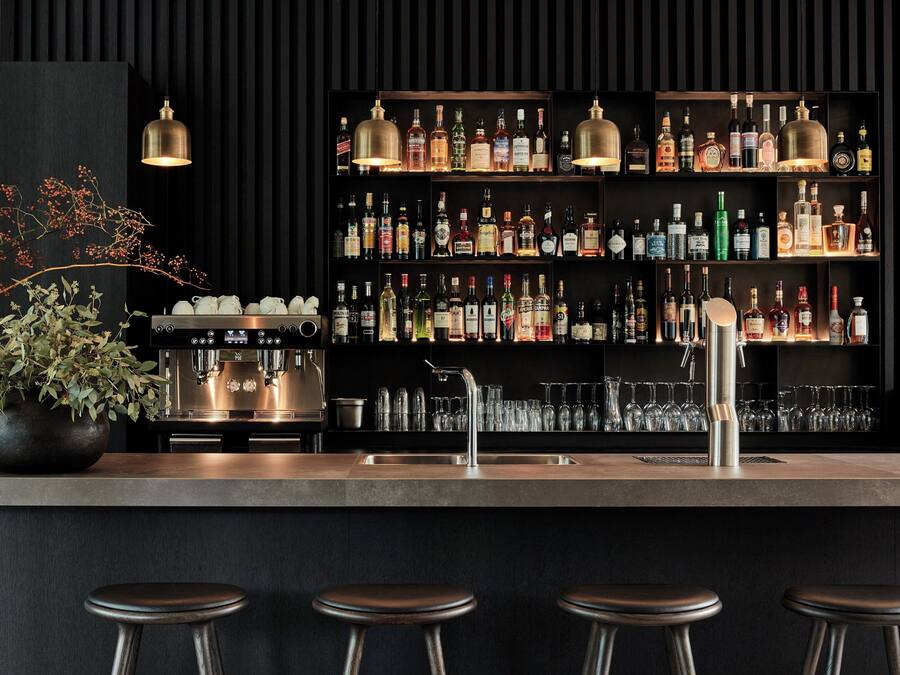 A bar area with a coffee machine, multiple shelves of liquor bottles, and bar stools.