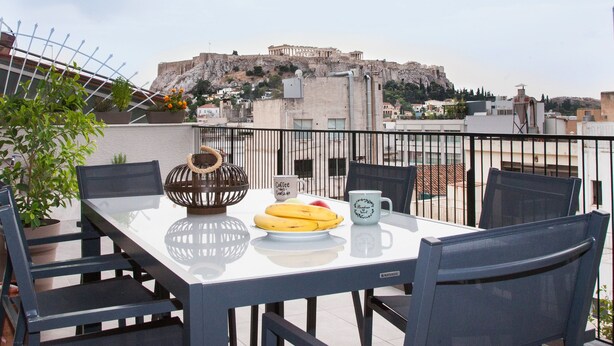 A balcony with a table set for two, featuring a plate of bananas, a coffee mug, and a decorative bowl. In the background, a historic building is visible on a hill. - Cozy Penthouse - Stunning View Acropolis (Athens)
