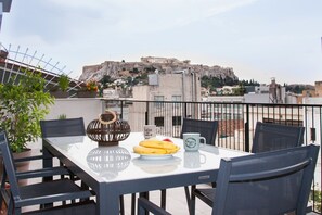 Een balkon met een tafel gedekt voor twee personen, met een bord bananen, een koffiemok en een decoratieve schaal. Op de achtergrond is een historisch gebouw op een heuvel te zien.