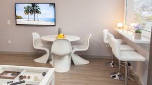 A modern room with a round table, white chairs, a TV displaying a beach scene, a small window with a view, and a bedside table with a lamp and a plant. - Cozy Penthouse - Stunning View Acropolis (Athens)