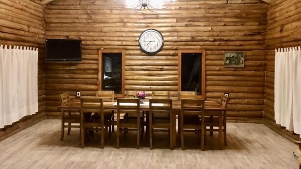 A wooden cabin interior with a dining table and chairs, a clock, a television, and two windows. - Cabaña La Emilia (Uspallata)