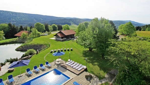 A pool area with lounge chairs and umbrellas, surrounded by a well-maintained lawn and a view of the mountains. - Berghotel Maibrunn (Sankt Englmar)