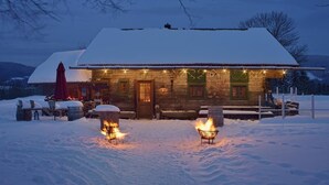 A log cabin with a snow-covered roof, a wooden door, and a small porch area with outdoor seating. - Berghotel Maibrunn (Sankt Englmar)
