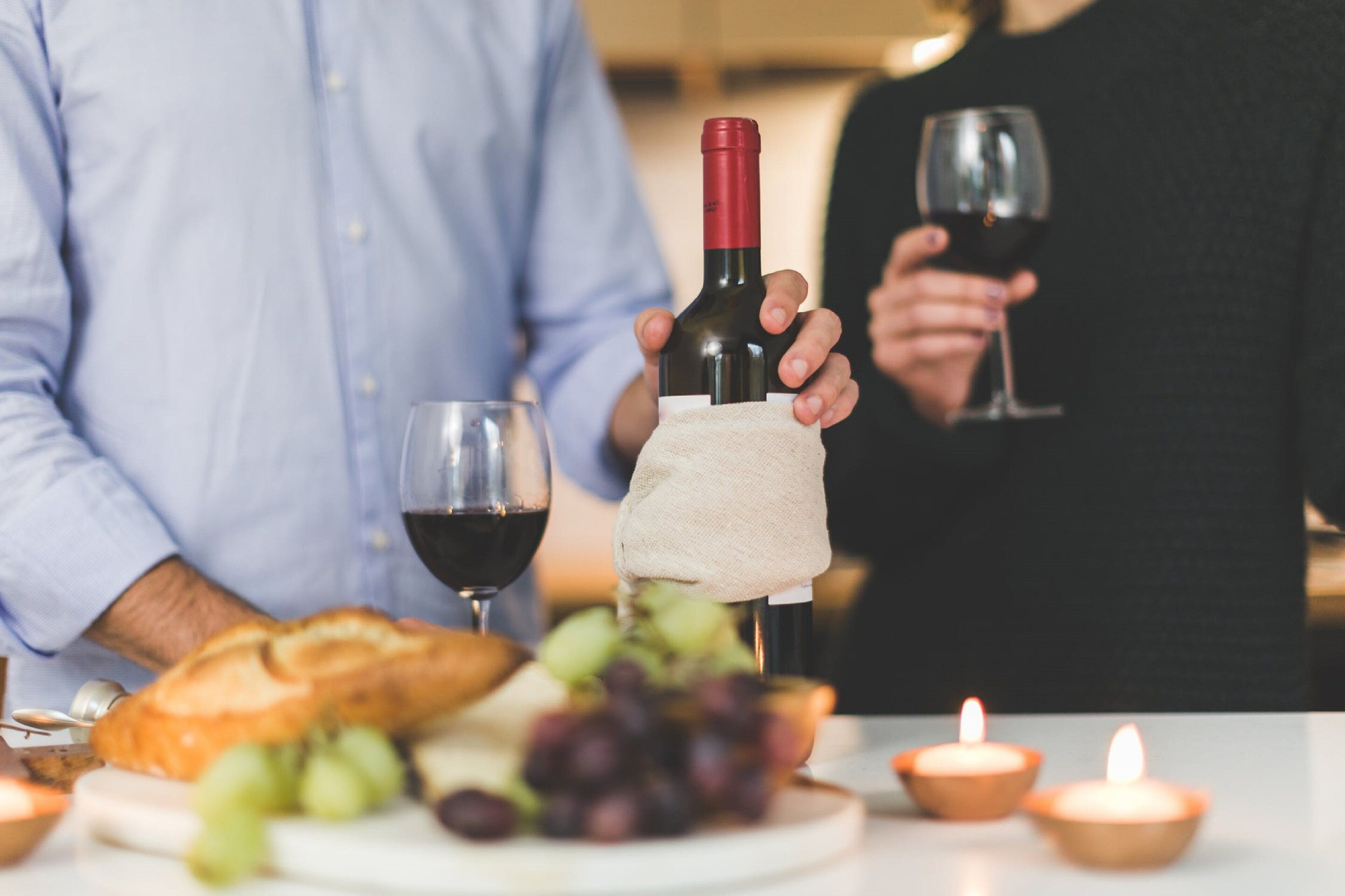 Una mesa con una botella de vino, copas, pan, uvas y velas.
