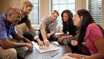 Un grupo de personas jugando un juego de mesa alrededor de una mesa redonda en una habitación con ventana y persianas.