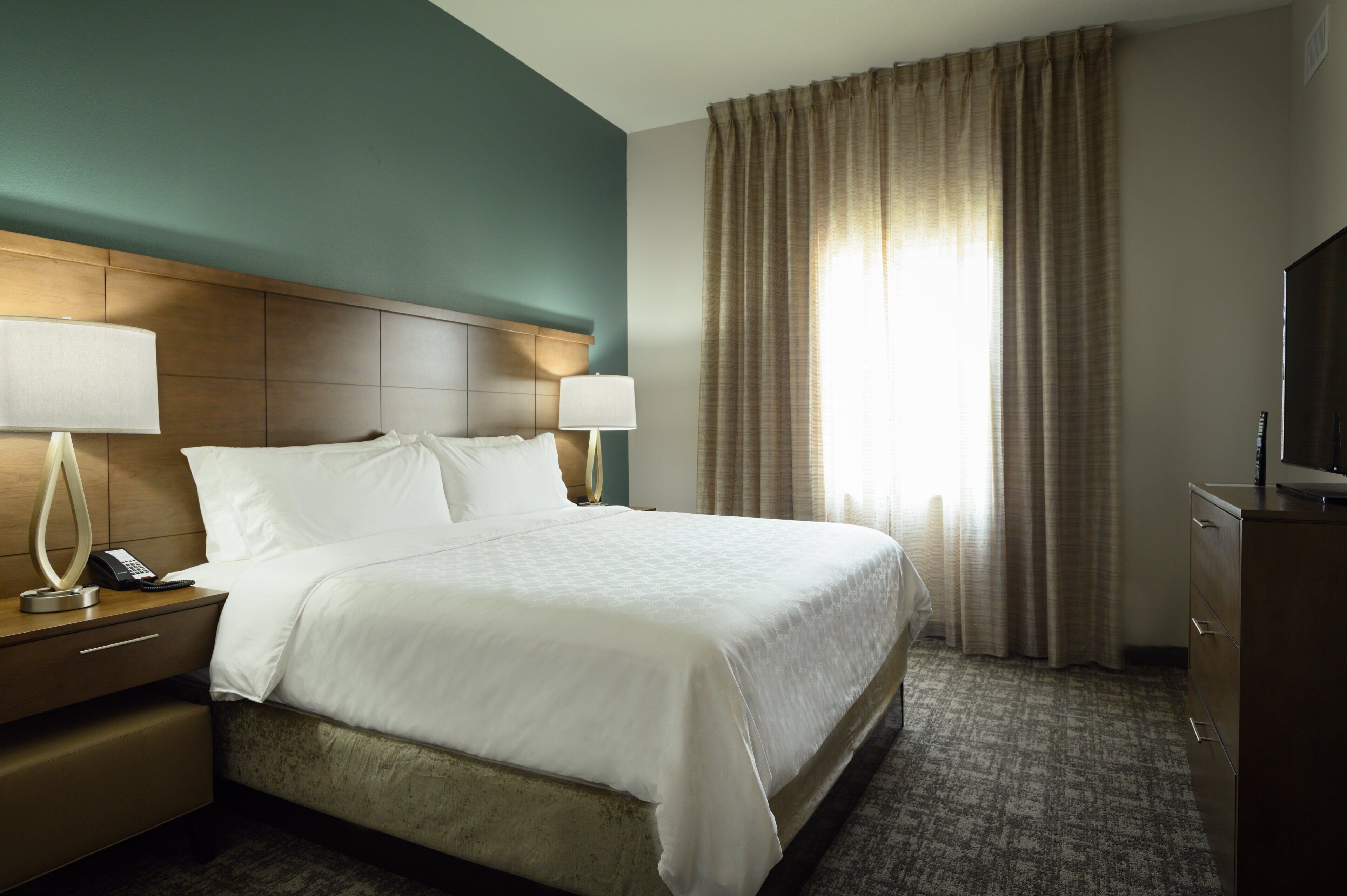 A hotel room with a large bed, two bedside tables with lamps, a television, and a window with curtains.