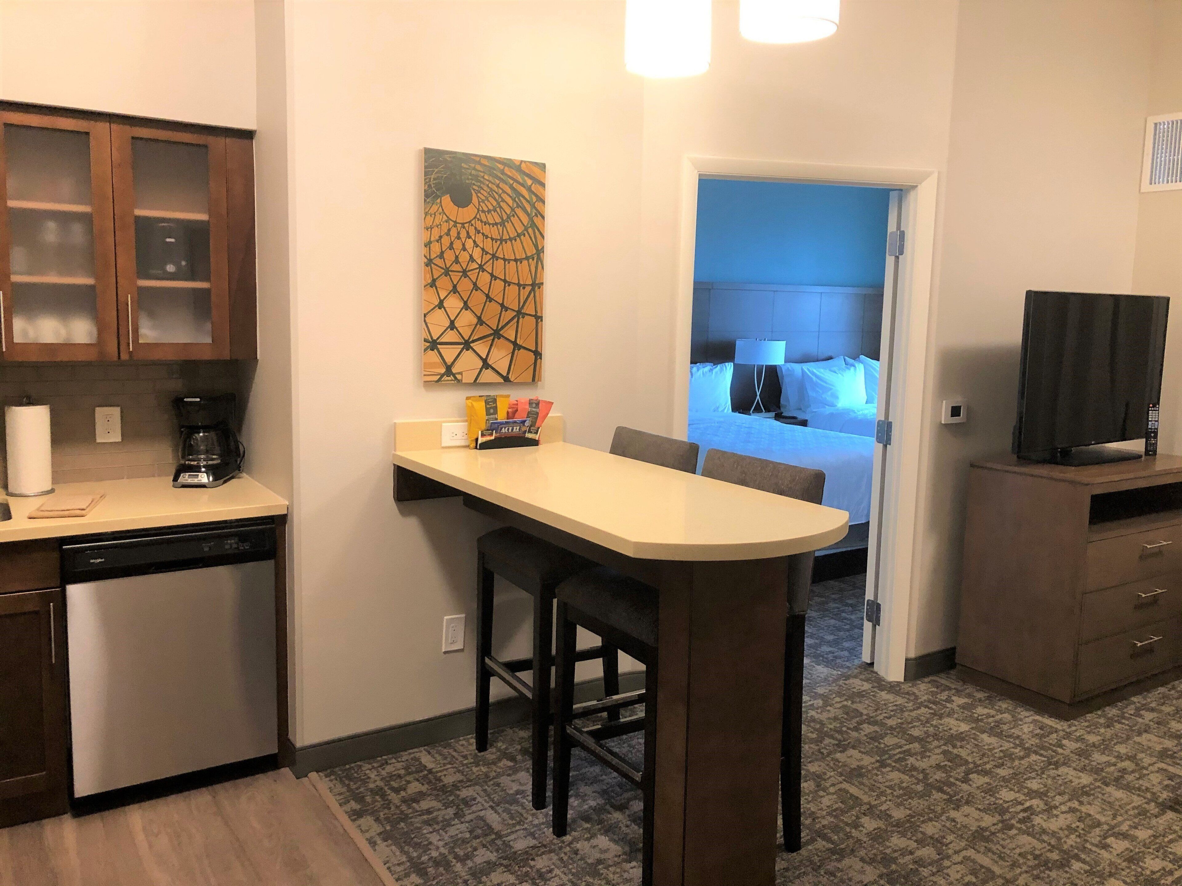 A compact kitchen with a coffee maker, a dining area with a small table and chairs, and a bedroom visible through an open door.