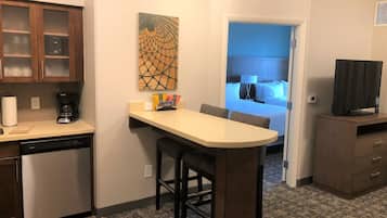 A compact kitchen with a coffee maker, a dining area with a small table and chairs, and a bedroom visible through an open door.
